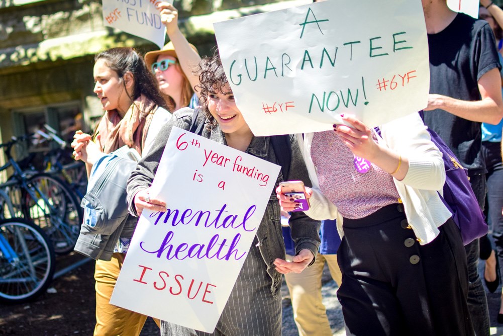 Two graduate workers hold up signs at a protest. One reads "6th year funding is a mental health issue." The other reads "A guarantee now!"