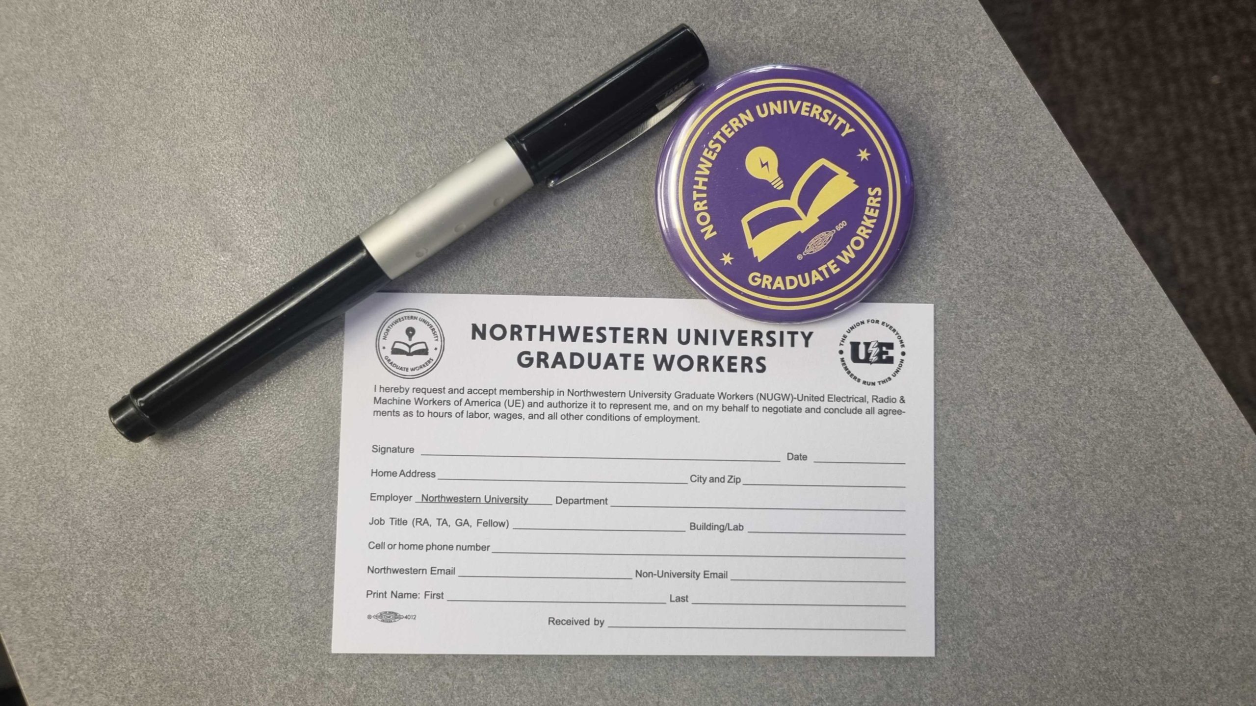 An NUGW union card sits next to a purple NUGW button and a pen, ready to be signed