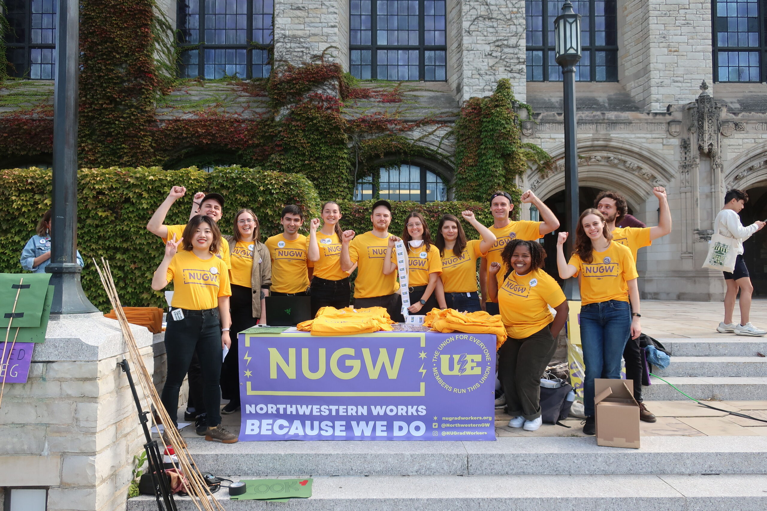 A group of 15 NUGW members stand in gold NUGW shirts in front of Deering Library. They are raising their fists in solidarity in front of an NUGW banner stating: NUGW, The University Works Because We Do!